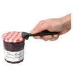 Brix JarKey Jar Opener Aluminium — small dark aluminium opener with curved nose held against the edge of a red-and-white checked lid on a Bonne Maman raspberry jam jar, a hand lifts the opener gently